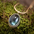 Bild in Galerie-Betrachter laden, Et nærbillede af *Vedhæng - Eryn Design - Tranquill 14kt Guld* viser en stor, oval-formet grønlig-blå ædelsten sat i en indviklet sølvring. Vedhænget er lavet af Eryn Design og hænger på en flettet 14 karat guldkæde og hviler på en seng af mos. Ædelstenen reflekterer lyset og giver den et lysende udseende.