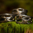 Bild in Galerie-Betrachter laden, En trio af mørke, teksturerede ringe fra Eryn Design-kollektionen, med et rustikt, organisk design, hviler oven på en seng af levende grønt mos. To af disse Fingerring - Moria (Simpel) ringe er håndlavede som håndlavede smykker i 925s Sterling Sølv og er prydet med små runde ædelsten sat i gyldne rammer, som tilføjer et elegant touch til den jordiske scene.