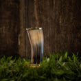 Bild in Galerie-Betrachter laden, Et poleret, buet Drikkeglas Horn (300 ml), med et skinnende ydre, står oprejst på en overflade omgivet af frisk grønt græs. Baggrunden har en rustik trævæg, der tilføjer en naturlig og jordnær stemning til billedet.
