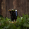 Bild in Galerie-Betrachter laden, Et poleret, buet Drikkeglas Horn (100 ml) med en messingkant hviler på en træoverflade omgivet af grønt løv. Baggrunden er sløret, hvilket fremhæver koppens håndværk og de naturlige omgivelser, der ligner et raffineret stykke drikkevare i sin elegance.