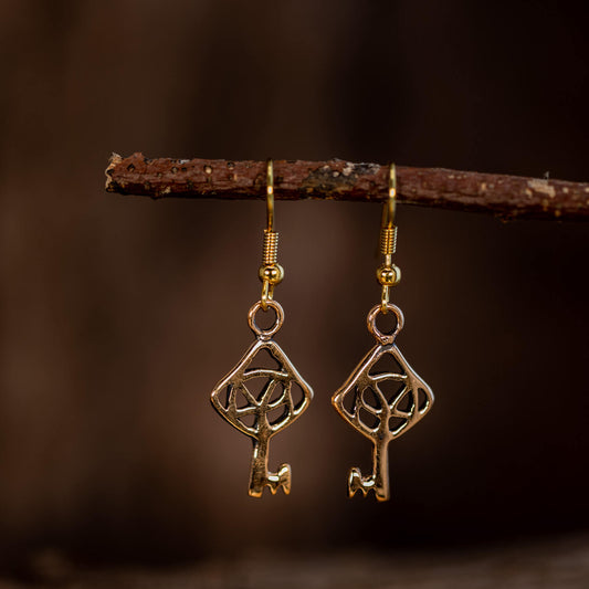 Et nærbillede af et par elegante Hængeøreringe Nøglen til Himmerriget Bronze hængende fra en gren. Øreringene, der er lavet i forgyldt sølv, har et geometrisk design, der ligner en nøgle med et indviklet trekantet og cirkulært mønster. Baggrunden er mørk og sløret, hvilket skaber en kontrast, der fremhæver de skinnende øreringe.