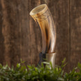 Bild in Galerie-Betrachter laden, Drikkehorn Rune, prydet med indviklede mønstre, er placeret på et træstativ mod en rustik træbaggrund. I forgrunden tilføjer grønt græs og planter et naturligt præg til scenen og fremkalder stemningen af vikingemarkeder.