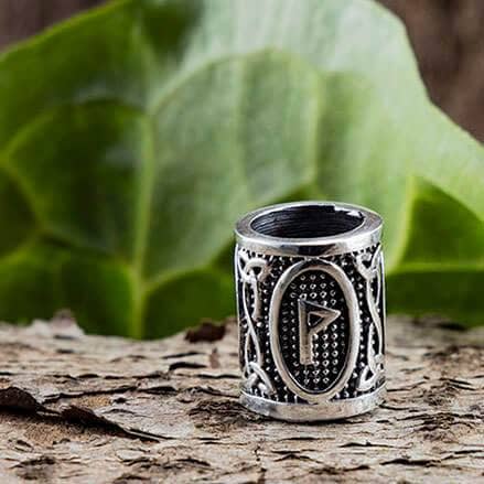 "Skægperler / Hårpynt Rune W Wunjo 925s Sølv", en sølvring med indviklede designs og en fremtrædende "W"-rune, der minder om Vikingesmykker, vises på en naturlig baggrund med store grønne blade og struktureret bark.