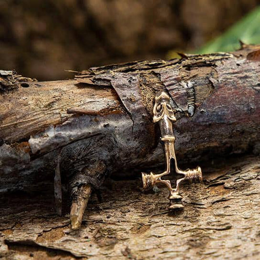 Et nærbillede af Islandsk Kors Vedhæng Bronze, et lille vedhæng, der er indviklet formet som et ø-kors, placeret på struktureret og forvitret træbark. Korset har detaljerede designs og små figurer i krydset. Den ru tekstur af barken i baggrunden tilføjer en rustik følelse til billedet.