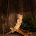 Bild in Galerie-Betrachter laden, En rustik, tekstureret baggrund viser Drikkehorn Raw m. Thors Hammer Holder hvilende på et træstativ. Hornet viser en naturlig gradient af beige og brune toner, der minder om traditionel vikinge-dekoration. Grønt løv ses i baggrunden, hvilket tilføjer et jordisk præg til indstillingen.