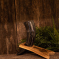 Bild in Galerie-Betrachter laden, Drikkehornet Ak m. Thors Hammer Holder står oprejst på en træbase med messingdetaljer, sat mod rustikke træplanker og frodigt grønt løv, der fremkalder essensen af vikingemarkeder.