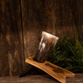 Bild in Galerie-Betrachter laden, Et poleret Drikkehorn Ak m. Thors Hammer Holder hviler på et træstativ mod en rustik træbaggrund. Bregneblade er placeret ved siden af stativet, hvilket tilføjer et strejf af grønt til scenen. Hornet har en glat, skinnende overflade med naturlige variationer i farve, der minder om genstande fundet på vikingemarkeder.