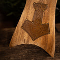 Bild in Galerie-Betrachter laden, Et nærbillede af et Drikkehorn Ak m. Thors Hammer Holder, med et detaljeret Thors hammer (Mjölnir) symbol fremhævet med messing søm på nøglepunkter. Træet har en varm tone og fyldig årer, der fremkalder det håndværk, man finder på vikingemarkeder. Baggrunden er blødt sløret med naturlige elementer.