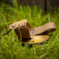 Bild in Galerie-Betrachter laden, En lille, brun lædersko med snørebånd delvist løsnet hviler på en rund træoverflade midt i grønt græs. I nærheden antyder et fint udformet Drikkehorn Wild historier om gamle traditioner. Den rustikke baggrund og bløde belysning tilføjer en naturlig, jordnær følelse til scenen.