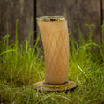 Bild in Galerie-Betrachter laden, Et "Drikkeglas Horn med mønster" (300ml) med guldkant og diamantformede graveringer, fyldt med en brun væske, står på en træskranke midt i højt grønt græs med en sløret træbaggrund.