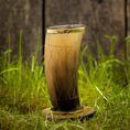 Bild in Galerie-Betrachter laden, Et dekorativt Drikkeglas Horn med mønster (300 ml), med en gradient fra mørk til lysebrun og prydet med ætsede geometriske mønstre, vises på en rund træstand midt i grønt græs med et sløret træhegn i baggrunden.