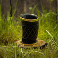 Bild in Galerie-Betrachter laden, Et Drikkeglas Horn med Mønster (100 ml), med et indviklet sort design med guldaccenter, er placeret på en træskranke omgivet af levende grønt græs. Baggrunden er let sløret, hvilket gør opmærksom på det unikke glass detaljerede mønster og skinnende finish.