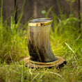 Bild in Galerie-Betrachter laden, Et Drikkeglas Horn med Mønster (100ml) med indviklede udskæringer og en gylden rand er placeret oprejst på en træskive, omgivet af frodigt grønt græs. Rammerne fremstår rustikke og naturlige.