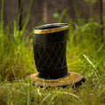 Bild in Galerie-Betrachter laden, Et sort Drikkeglas Horn med Mønster (100 ml), indviklet designet med guldkant, hviler på et lille, rundt stykke træ i græsset. Baggrunden viser sløret grønt løv og en utydelig træstruktur, der fremkalder charmen og det unikke ved håndværksfremstillede stykker.