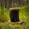 Bild in Galerie-Betrachter laden, Et poleret, mørkt Drikkeglas Horn med Mønster (100ml) med en gylden kant sidder oven på en træbase på græs. Koppen har et krydsmønster ætset ind i overfladen og er sat mod en sløret baggrund med naturlige elementer.