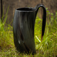 Bild in Galerie-Betrachter laden, Et Vikingekrus Nature Horn (600-800 ml), med et slankt, mørkt, buet design med et håndtag, står på en plet græs. Kruset har en poleret, skinnende overflade og en organisk let snoet form. Baggrunden er lidt sløret og ser ud til at være af træ.