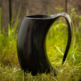 Bild in Galerie-Betrachter laden, Et sort Vikingekrus Nature Horn (600-800ml) med et håndtag sidder oprejst på grønt græs, med et træhegn sløret i baggrunden. Hornet, der ligner traditionelle vikingekopper, har en blank finish og buer i en unik form.