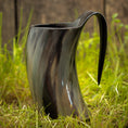 Bild in Galerie-Betrachter laden, Et poleret, buet Vikingekrus Nature Horn (600-800ml) med et håndtag, stående på græsklædt jord med en sløret træbaggrund. Dette sorte drikkehorn har en blank finish og naturlige teksturerede mønstre, hvilket giver et unikt og rustikt udseende.