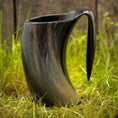 Bild in Galerie-Betrachter laden, Et sort Vikingekrus Nature Horn (600-800ml) med en hornlignende form sidder på en græsklædt overflade. Denne unikke håndlavede krus har en blank finish, en bred åbning og et buet håndtag, der ligner et horn. Baggrunden er sløret, hvilket understreger kruset som fokus.