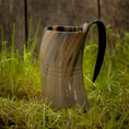 Bild in Galerie-Betrachter laden, Et poleret, hornformet Vikingekrus Helleristning Horn (600-800ml) med et håndtag sidder på frodigt grønt græs. Baggrunden er en rustik trævæg, der forstærker scenens naturlige og historiske fornemmelse. Håndlavede hornkrus har subtile linjer og varierende nuancer af brun, tan og sort.