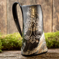 Bild in Galerie-Betrachter laden, Vikingekrus Yggdrasil Horn, med et mørkt hornformet design med et hvidt keltisk knudemønster, der ligner Yggdrasil ask indgraveret på dets forside, hviler på en rustik træoverflade med en mosbeklædt baggrund, hvilket skaber en naturlig og jordnær stemning. Dette unikke krus har en rigelig kapacitet på 600-800ml.