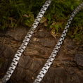 Bild in Galerie-Betrachter laden, Nærbillede af to Halskæde Panzering 5,5 mm 925s Sølv-kæder lagt parallelt på et stykke struktureret, barkbeklædt træ. Grønt mos omgiver halskæden og tilføjer et naturligt element til scenen. Kæderne har et zigzag-mønster og et geometrisk, sammenhængende design i kontrast til den organiske baggrund.