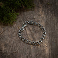 Bild in Galerie-Betrachter laden, Armbånd Chainlinked 5mm 925s Sølv, lavet af oxideret 925s Sterling sølv, ligger på en forvitret træoverflade. Noget grønt mos er synligt i det øverste venstre hjørne af billedet, hvilket tilføjer et naturligt touch til den rustikke baggrund og fremhæver denne unisex-smykkes rå elegance.