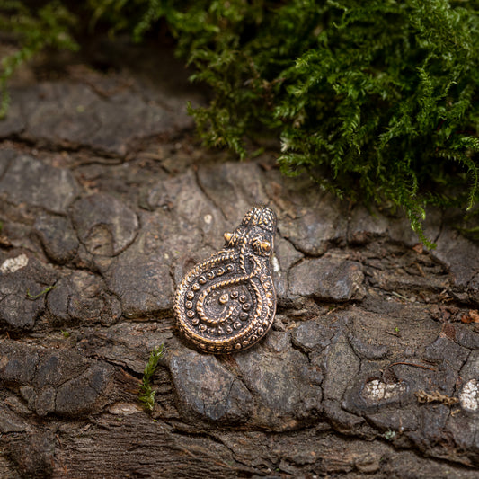 Vedhæng Slangen fra Gørding Bronze, en detaljeret metallisk amulet med et indviklet firbendesign, sidder på bark med ru tekstur, omgivet af grønt mos.