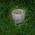 Bild in Galerie-Betrachter laden, En opretstående Lysestage til fyrfadslys, cylindrisk og gennemskinnelig, placeret på en plet af grønt græs. Lignet til en delikat messingholder virker den tom og har en glat overflade med en let glans.