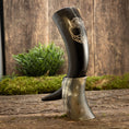 Bild in Galerie-Betrachter laden, To udsøgte Drikkehorn Yggdrasil U. Mønster, med den ene i et graveret sort design og den anden i en naturlig hornnuance, fremvises stående på en struktureret træoverflade prydet med grønt mos og planter som baggrund.