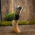 Bild in Galerie-Betrachter laden, Drikkehorn Templar, et dekorativt vikinge-drikkehorn lavet af polerede bøfler med indviklede udskæringer, hviler oprejst på et træstativ. Baggrunden har en rustik træoverflade prydet med grønt mos og løv, der forstærker den naturlige og historiske æstetik, der minder om vikingemarkeder.