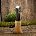 Bild in Galerie-Betrachter laden, En poleret Drikkehorn Templar står oprejst i en beige meleret holder på en rustik træoverflade. Frisk grønt løv og mos fremhæver de jordiske omgivelser, og hornet har et indviklet udskåret design nær toppen, der minder om dem, man finder på vikingemarkeder.