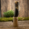 Bild in Galerie-Betrachter laden, En poleret Drikkehorn Templar med gyldne graveringer hviler i et indviklet formet hornstativ. Opsætningen er placeret på en rustik træflade med en plet grønt og en trævæg i baggrunden, hvilket skaber en middelalderlig æstetik, der minder om vikingemarkeder.