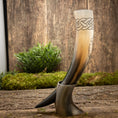 Bild in Galerie-Betrachter laden, Drikkehorn Yggdrasil med Mønster, prydet med indviklede keltiske knudedesigns øverst, vises opretstående på en rustik træoverflade ved hjælp af et stativ. Grønt løv og mos i baggrunden skaber naturlige, jordnære omgivelser, der minder om et vikingemarked.