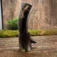 Bild in Galerie-Betrachter laden, En poleret Drikkehorn Valknut med indviklede udskæringer står oprejst på en robust, forvitret træoverflade. Baggrunden har en rustik trævæg og grønt mos, der fremkalder vikingetidens naturlige, jordiske atmosfære.