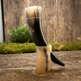 Bild in Galerie-Betrachter laden, Drikkehorn Valknut, med udsøgt sort og hvid marmorering og ætset med et slående geometrisk design, hviler elegant på en naturholder træstativ. En mosbeklædt jord og træbaggrund forstærker dens naturlige, historiske charme yderligere, mens en lille bladplante tilføjer et strejf af grønt i baggrunden.
