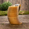 Bild in Galerie-Betrachter laden, Et nærbillede af Vikingekrus Bark Horn (600-800ml) sat på en rustik træoverflade med grønt mos og planter i baggrunden. Ølkrusen viser en jævn gradient fra lys til mørk og har lodrette riller langs den øverste halvdel.