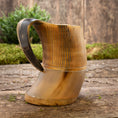 Bild in Galerie-Betrachter laden, Et Vikingekrus Bark Horn (600-800ml) med træbund og håndtag hviler på en rustik træoverflade. Hornets krop er poleret med en naturlig gradient af lyse til mørkebrune nuancer, med udskårne lodrette linjer nær toppen. Grønne buske er slørede i baggrunden.