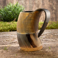 Bild in Galerie-Betrachter laden, Et rustikt, håndlavet Vikingekrus Bark Horn (600-800ml) med lodrette kanter på den øverste halvdel og et glat, buet håndtag sidder på en træoverflade. I baggrunden er der grønt og en trævæg, der tilføjer en naturlig, jordnær følelse til scenen.