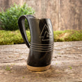 Bild in Galerie-Betrachter laden, Et Vikingekrus Valknut Horn (600-800ml), sort og minder om vikingetraditioner, sidder på en træbase på en rustik træoverflade. Hornet er indgraveret med et trekantet design og tre vandrette linjer. I baggrunden er der grønt og en trævæg, der forstærker den naturlige og rustikke stemning.
