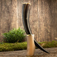 Bild in Galerie-Betrachter laden, Drikkehorn Ordained Galore, et elegant aflangt bøffelhorn prydet med metalaccenter, står majestætisk på en rustik træoverflade dækket af grønt mos. Baggrunden har en tekstureret trævæg, mens en nærliggende lille busk introducerer yderligere grønt til de jordnære omgivelser. De indviklede metaldetaljer vækker minder om skatte fra vikinge-marker.