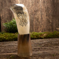 Bild in Galerie-Betrachter laden, Drikkehorn Valravn, udsmykket med et indviklet design af to sammenflettede fugle, står oprejst på en træflade omgivet af mos og grønt. Bagtæppet er en tekstureret trævæg, der fremkalder en rustik og naturlig stemning, der minder om et vikingemarked.