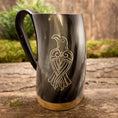 Bild in Galerie-Betrachter laden, Et poleret Vikingekrus Valravnen Horn (600-800 ml) har et indgraveret design af en stiliseret fugl på siden. Tankarden viser et sort håndtag og en metallisk base, der hviler elegant på en rustik træoverflade med grønt mos i baggrunden.