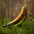 Bild in Galerie-Betrachter laden, En poleret, buet Drikkehorn Devine med udsmykkede metalbånd i begge ender hviler på en plet af grønt græs. Hornet, der minder om dem, man fandt på vikingemarkeder, er støttet op med en sort rem, og en rustik trækulisse ses i baggrunden.