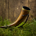 Bild in Galerie-Betrachter laden, En Drikkehorn Devine, med en udsmykket messingbeklædning og en sort rem, ligger poleret på en græsklædt overflade på baggrund af et træhegn, der minder om skatte fundet på vikingemarkeder.