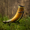 Bild in Galerie-Betrachter laden, En Drikkehorn Devine med elegante metalaccenter hviler på en græsklædt overflade. Hornet er placeret oprejst og understøttet af et sort metalstativ, sat mod den rustikke kulisse af en trævæg, der minder om scener fra vikingemarkeder.