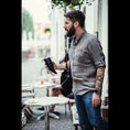 Bild in Galerie-Betrachter laden, En skægget mand iført en grå skjorte og jeans står udenfor en café med en Gede Kaffe Drikkehorn Sort. Han bærer en sort skuldertaske og har et tatoveringsærme på venstre arm. Der er tomme borde og stole omkring ham, med slørede individer i baggrunden.