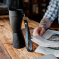 Bild in Galerie-Betrachter laden, En person i en ternet skjorte holder en avis med den ene hånd, mens en sort ged Kaffe Drikkehorn Sort lavet af BPA-fri plast og en læder nøglering med teksten "GOAT STORY" er placeret på bordet foran dem. Baggrunden er lidt sløret.