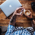 Bild in Galerie-Betrachter laden, En person med tatoveringer på underarmen, iført en ternet skjorte, sidder ved et træbord. De holder en kop med latte art i den ene hånd og åbner låget på deres Ged Kaffe Drikkehorn Brun med den anden. En bærbar computer og et kamera er også på bordet, og fuldender scenen for denne moderne viking.