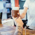 Bild in Galerie-Betrachter laden, En tatoveret arm rækker en hånd ud for at tage imod en ged Kaffe Drikkehorn Brun fra en barista på tværs af cafedisken, der minder om en moderne viking, der handler med sit drikkehorn. Baggrunden er sløret og fokuserer scenen på kaffeoverdragelsen.